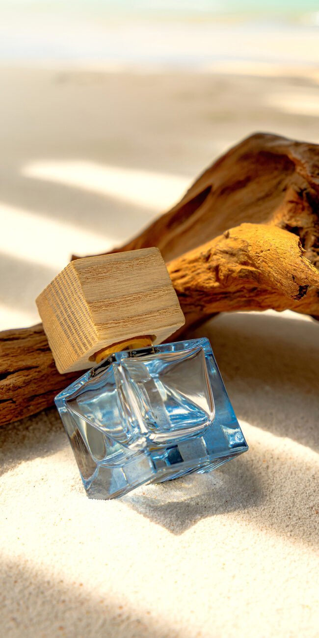 Bouteille de parfum bleu avec bouchon en bois posée sur du sable à côté d'une branche de bois flotté, capturée par Studio Helloday.