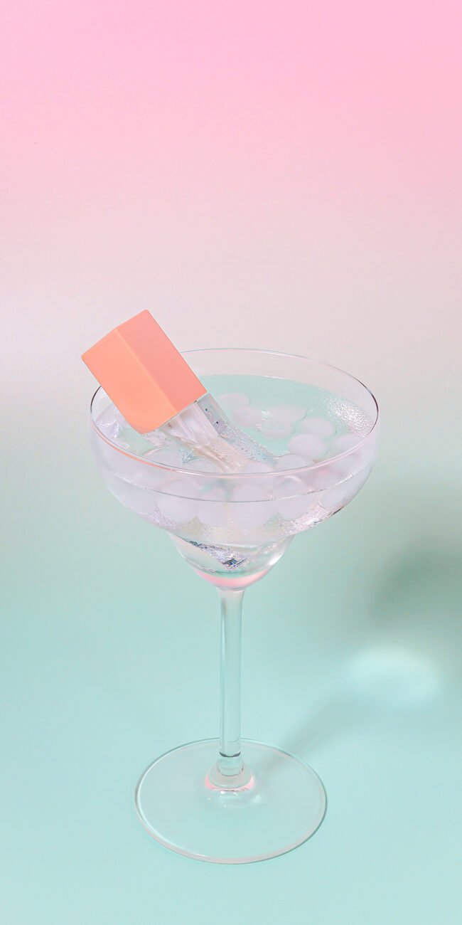 Gloss cosmétique transparent dans un verre avec des glaçons sur fond pastel rose et vert, photographié par Studio Helloday