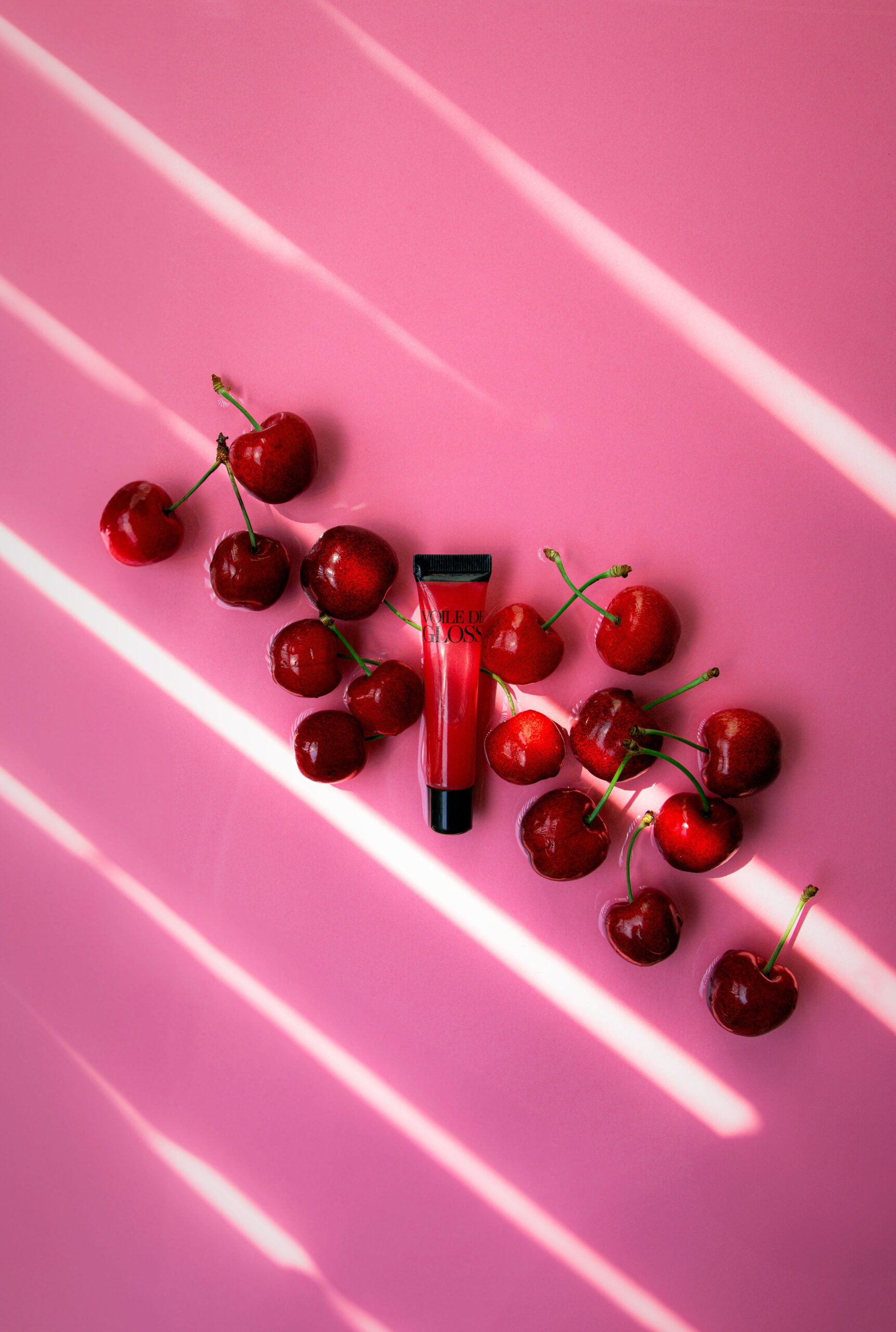 Photo produit Studio Helloday : gloss rouge entouré de cerises sur fond rose avec lumière en raies.
